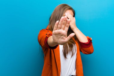 Genç bir kadın yüzünü eliyle kapatıyor ve kamerayı durdurmak için diğer elini öne koyuyor. Mavi arka planda fotoğraf veya resimleri reddediyor.