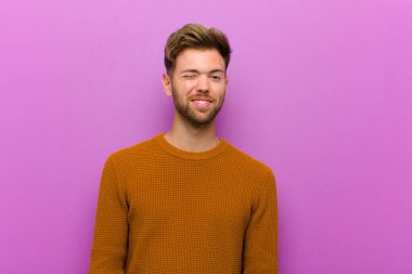 Genç adam mutlu ve arkadaş canlısı görünüyor, gülümsüyor ve mor arka plana karşı olumlu bir tavırla sana göz kırpıyor.