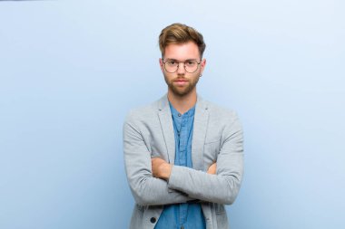 Genç işadamı mutsuz ve hayal kırıklığına uğramış hissediyor. Ciddi görünüyor. Mavi arka planda çapraz kollara kızgın ve kızgın.