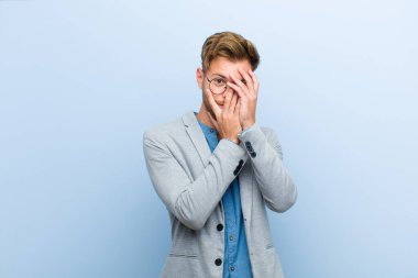 Genç işadamı korkmuş ya da utanmış hissediyor. Mavi arka planda elleri yarı kapalı bir şekilde dikizliyor ya da dikizliyor.