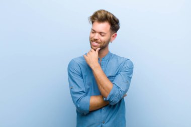 young businessman smiling with a happy, confident expression with hand on chin, wondering and looking to the side against blue background