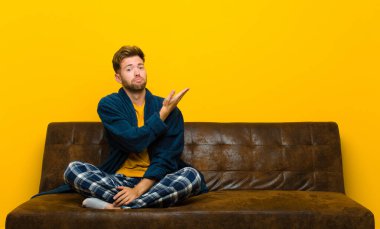 Pijama giyen genç adam kafası karışık ve habersiz, şüpheli bir açıklama ya da düşünceyi merak ediyor. Kanepede oturuyor.