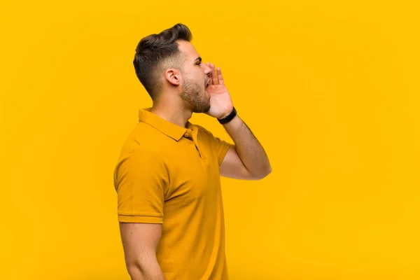 young hispanic man profile view, looking happy and excited, shouting and calling to copy space on the side against orange wall