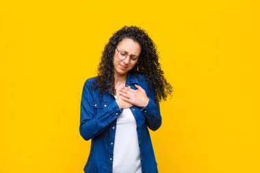 Genç, güzel bir kadın üzgün, incinmiş ve kalbi kırık görünüyor. İki elini de kalbine yakın tutuyor. Ağlıyor ve turuncu duvara yaslanıyor.