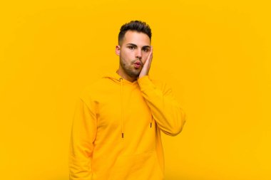 Genç İspanyol adam şok olmuş ve şok olmuş hissediyor. Ağzı açık halde turuncu duvara yaslanmış, yüz yüze inanamıyor.