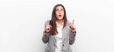 Genç, güzel bir kadın, şaşkınlık ve şaşkınlık içinde, ağzı açık bir şekilde beyaz duvara yaslanmış, şaşırmış bir şekilde yukarı bakıyor.