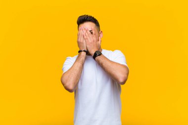 Genç İspanyol adam elleriyle yüzünü kapatıyor. Şaşırmış bir ifadeyle parmaklarının arasından bakıyor ve turuncu duvara bakıyor.