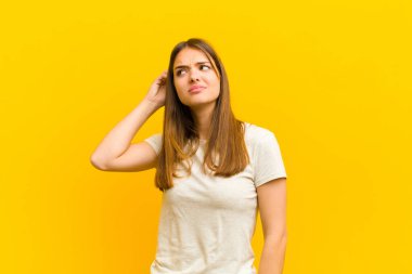 Genç güzel kadın şaşkın ve kafası karışmış hissediyor, başını kaşıyor ve turuncu arka plana bakıyor.