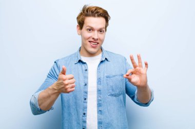 Genç kızıl saçlı adam kendini mutlu, şaşkın, tatmin olmuş ve şaşırmış hissediyor, tamam gösteriyor ve işaret ediyor, mavi duvara gülümsüyor.