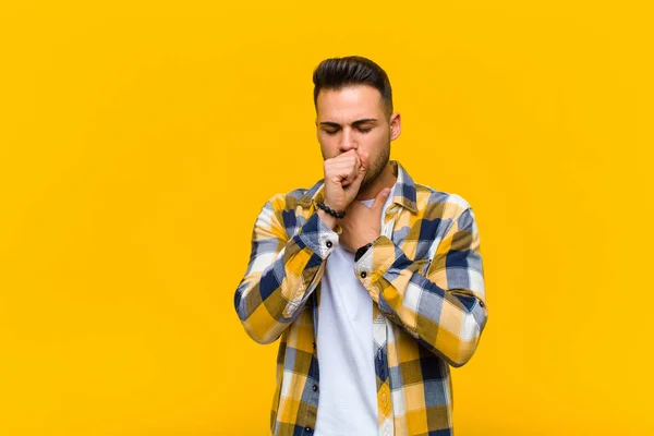 young hispanic man feeling ill with a sore throat and flu symptoms, coughing with mouth covered against orange wall