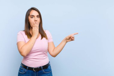Genç, güzel bir kadın kendini mutlu, şok olmuş ve şaşırmış hissediyor. Ağzını eliyle kapatıyor ve mavi arka plana karşı yanal fotokopi alanını işaret ediyor.