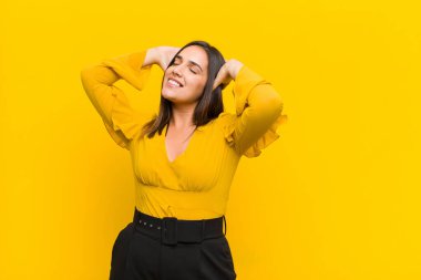 Genç ve güzel bir kadın gülümsüyor ve rahatlıyor, tatmin oluyor ve kaygısız, gülüyor ve turuncu duvarda izole bir şekilde dinleniyor.