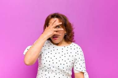 Orta yaşlı bir kadın şok olmuş, korkmuş ya da korkmuş görünüyor. Yüzü elle örtülmüş ve parmaklarının arasından mor duvara bakıyor.