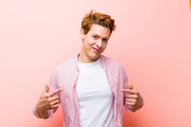 Genç kızıl saçlı adam gururlu, pozitif ve rahat görünüyor. İki eli de pembe duvara yaslanmış göğsünü gösteriyor.
