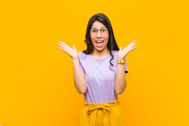 Genç ve güzel Latin kadın mutlu ve heyecanlı görünüyor. Beklenmedik bir sürprizle şoke olmuş durumda. İki eli turuncu duvarın yanında.