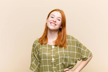 Genç kızıl saçlı güzel bir kadın mutlu bir şekilde gülümsüyor, eli kalçasında ve kendinden emin, olumlu, gururlu ve arkadaş canlısı bir tavırla.