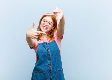 Genç kızıl saçlı güzel bir kadın mutlu, arkadaş canlısı ve pozitif hissediyor, gülümsüyor ve elleriyle bir portre veya fotoğraf çerçevesi yapıyor.