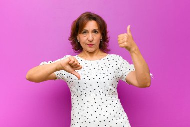 Orta yaşlı bir kadın, kafası karışık, bilgisiz ve kararsız hissediyor, iyi ve kötü seçenekleri değerlendiriyor veya mor duvara karşı seçenekleri değerlendiriyor.