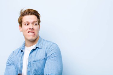 Genç kızıl saçlı adam şüphe ediyor ya da düşünüyor, dudağını ısırıyor ve kendini güvensiz ve gergin hissediyor, mavi duvara karşı boşluğu kopyalamak istiyor.