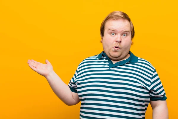 young big size man looking surprised and shocked, with jaw dropped holding an object with an open hand on the side against orange wall