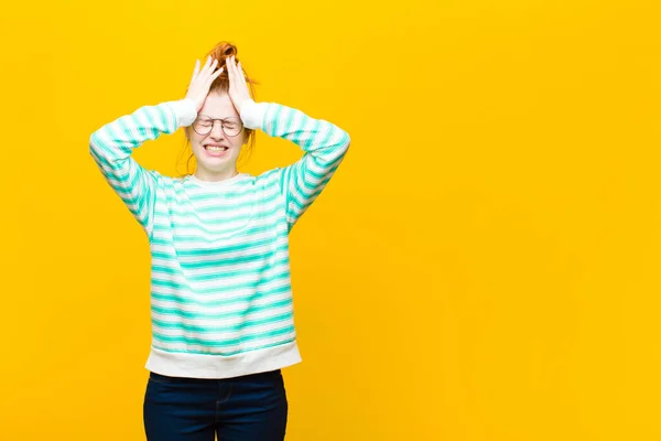 young red head woman feeling stressed and anxious, depressed and frustrated with a headache, raising both hands to head against orange wall