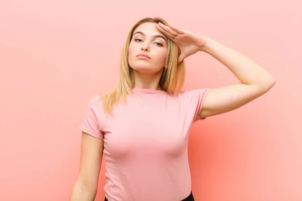 young pretty blonde woman greeting the camera with a military salute in an act of honor and patriotism, showing respect against pink flat wall