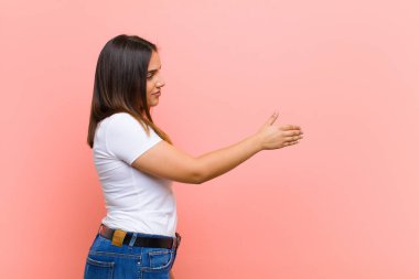 Genç, güzel İspanyol kadın neşeyle gülümsüyor sıcak, dostça, sevgi dolu bir hoş geldin sarılması yapıyor, pembe duvara karşı mutlu ve sevimli hissediyor.