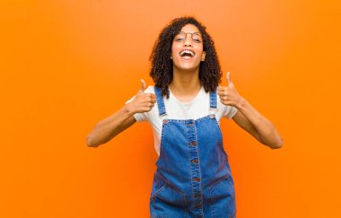 young pretty black woman smiling broadly looking happy, positive, confident and successful, with both thumbs up against orange wall clipart
