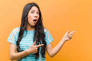 Genç, güzel, İspanyol bir kadın şok olmuş ve şaşırmış hissediyor. Yan taraftaki fotokopi alanını işaret ediyor. Kahverengi duvara karşı şaşkın, açık ağızlı bir bakış.