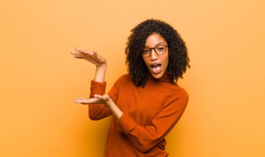 Genç, güzel, siyah bir kadın, iki eli de yanda olan bir objeyi tutuyor. Fotokopi alanı, turuncu duvara karşı bir objeyi gösteriyor, sunuyor ya da reklamını yapıyor.