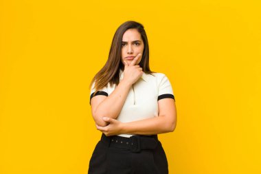 Genç, güzel bir kadın ciddi, düşünceli ve güvensiz görünüyor, bir kolu çapraz ve çene üzerinde, turuncu duvarda izole seçenekleri tartıyor