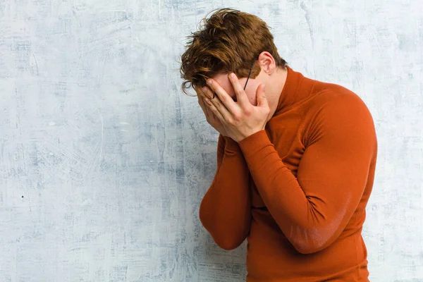 young red head man covering eyes with hands with a sad, frustrated look of despair, crying, side view against grunge wall