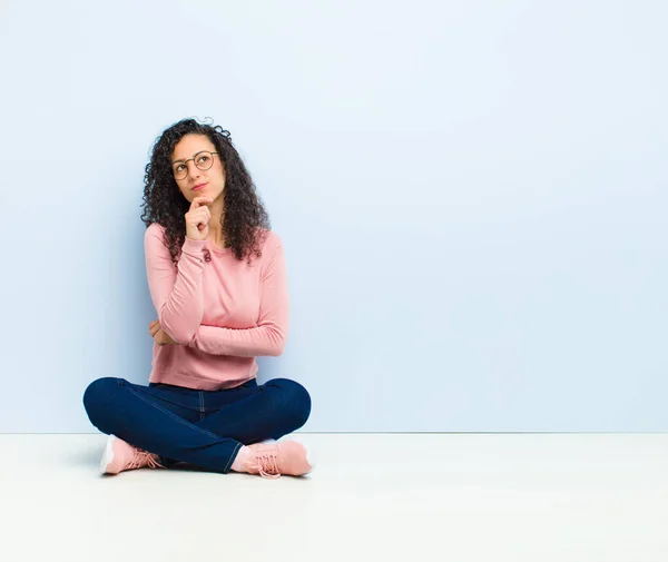 young pretty woman feeling thoughtful, wondering or imagining ideas, daydreaming and looking up to copy space sitting on floor