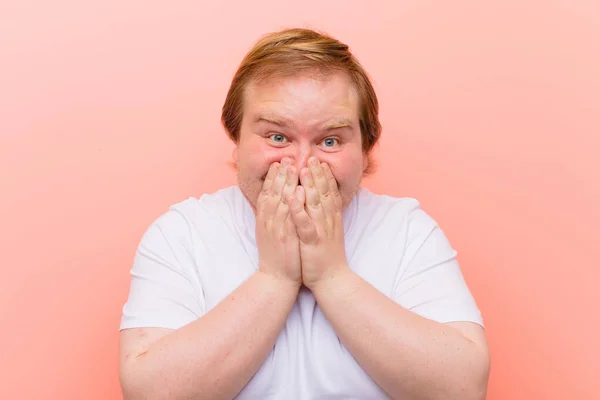 young big size man looking happy, cheerful, lucky and surprised covering mouth with both hands against pink wall