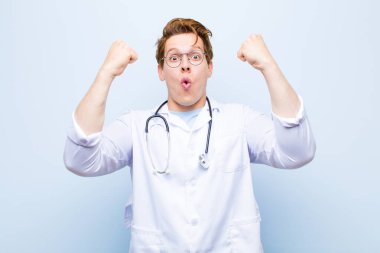 Genç kızıl saçlı doktor inanılmaz bir başarıyı kutluyor. Kazanan gibi. Heyecanlı ve mutlu görünüyor. mavi duvara yaslan