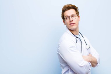 Genç kızıl saçlı doktor neşeyle gülümsüyor, mutlu, tatmin olmuş ve rahatlamış hissediyor, kollarını kavuşturup mavi duvara bakıyor.