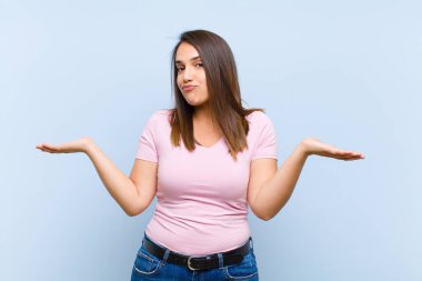 Genç, güzel bir kadın şaşkın ve şaşkın, şüpheci, farklı seçenekleri tartıyor ya da seçiyor. Mavi arka planda komik bir ifade var.