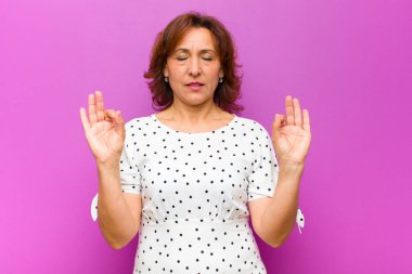 Orta yaşlı kadın konsantre olmuş ve meditasyon yapıyor, tatmin olmuş ve rahatlamış hissediyor, düşünüyor ya da mor duvara karşı bir seçim yapıyor.