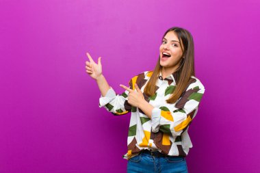 Genç, güzel bir kadın sevinçli ve şaşırmış hissediyor, şok olmuş bir ifadeyle gülümsüyor ve mor arkaplanı işaret ediyor.