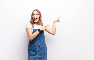 Genç, güzel bir kadın sevinçli ve şaşırmış hissediyor. Şok olmuş bir ifadeyle gülümsüyor ve beyaz duvara doğru işaret ediyor.