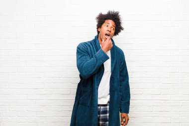 Önlüklü genç siyah adam şaşırmış, gergin, endişeli ya da korkmuş görünüm ile pijama giyiyor, duvara karşı fotokopi odasına doğru bakıyor.