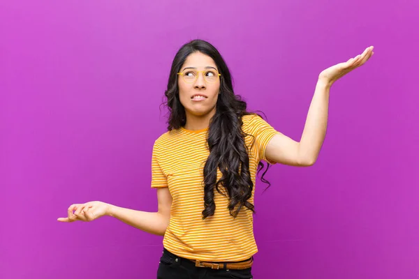 young pretty latin woman shrugging with a dumb, crazy, confused, puzzled expression, feeling annoyed and clueless against purple wall