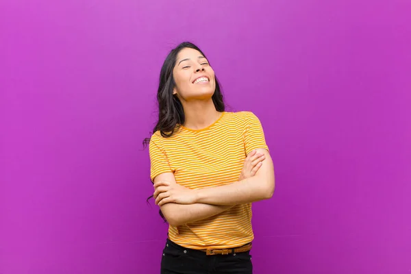 young pretty latin woman laughing happily with arms crossed, with a relaxed, positive and satisfied pose against purple wall