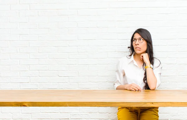 young  pretty latin woman doubting or thinking, biting lip and feeling insecure and nervous, looking to copy space on the side sitting in front of a table