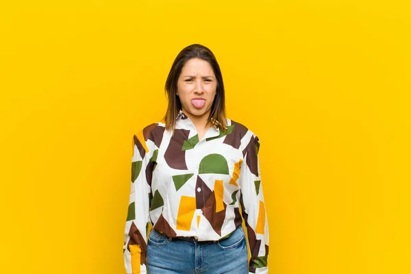 latin american woman feeling disgusted and irritated, sticking tongue out, disliking something nasty and yucky isolated against yellow wall