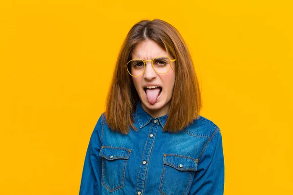 young pretty woman feeling disgusted and irritated, sticking tongue out, disliking something nasty and yucky against yellow background