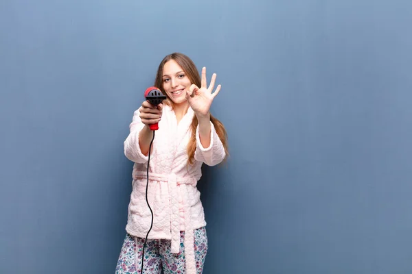 young pretty woman wearing pajamas and holding a hairdresser against blue wall with a copy space