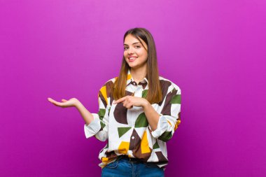 Genç ve güzel bir kadın neşeyle gülümsüyor ve avucunun kenarındaki fotokopi alanını işaret ediyor, mor arka plana karşı bir nesneyi gösteriyor veya reklamını yapıyor.