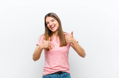 Genç ve güzel bir kadın gülümsüyor. Mutlu, pozitif, kendinden emin ve başarılı görünüyor. İki başparmağı da beyaz arka planda.