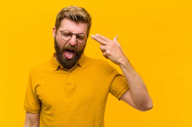 Genç sarışın adam mutsuz ve stresli görünüyor, intihar jesti elle silah işareti yapıyor, kafasını turuncu duvara doğrultuyor.
